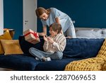 Young woman loving mother surprising son with birthday gift at home, mom giving wrapped box to excited little boy, congratulating child. Overjoyed happy kid sitting on sofa getting present from mum