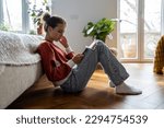 Small photo of Teens and cyberbullying. Upset teen girl sitting on floor near bed using smartphone at home, scrolling social media. Child spending too much time on phone. Teenagers and gadget addiction