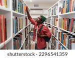 Small photo of Nerd student african american man choosing book in university library taking it from bookshelf. Wistful guy hipster with backpack studying college. Education, giving new profession, learning concept.