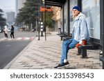 Young blond guy student waiting transport very long time in morning.Man on bus stop with coffee away. Blonde hair millennial man on bus stop in city. Modern urban transport for everyone