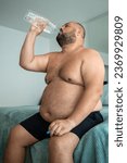 Small photo of Extremely fat guy drinks lot of water, sits on bed, trying to cool off, alleviate serious condition, resist summer heat. Chubby, over obese, oversize man copes with dehydration due to profuse sweating