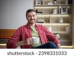 Small photo of Positive joyful carefree middle-aged bearded man with encouraging sympathetic smile. Cheerful male sitting at home, holding cup, drinking tea, looking at camera friendly smiling showing teeth