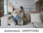 Sad tired woman sits on couch at home wrapped in plaid in living room. Blue mood of depressed girl boring. Nervous female feeling frustration hiding from society in loneliness solitude. Life problems.