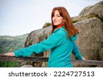 Small photo of Beautiful young smiling woman tourist in blue fleece sweater and orange scarf, with red hair standing in tourist place in mountains, enjoying the view