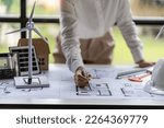 Small photo of Female engineer points to blueprints for house hoist and wind turbine next to concept of building house with renewable energy installation with wind turbine and solar cells