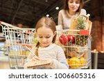 Warm-toned portrait of family doing grocery shopping in supermarket, focus on little girl holding shopping list standing by cart