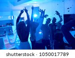 Group of modern young people dancing under confetti at private house party lit by blue light