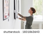 Small photo of Side view portrait of tattooed creative woman hanging paintings on wall while planning art gallery exhibition, copy space