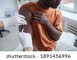 Small photo of Close up of adult African-American man looking away while getting covid vaccine in clinic or hospital, with male nurse injecting vaccine into shoulder