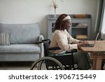 Side view portrait of young African-American woman using wheelchair while working from home in minimal grey interior, copy space