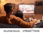 Back view of adult couple watching TV at home while sitting on sofa lit by warm cozy light, copy space