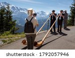 Small photo of Schynige Platte, Bernese Oberland, Switzerland - August 1 2019 : 2 Alpenhorn players in traditional swiss outfit filmed by female tourist on mobile phone. Alpine panorama background