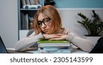 Small photo of Close up of preteen girl sitting by table and thinking about solving school lesson exams among a large pile of books with sad face on background bookcase at home