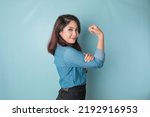 Excited Asian woman wearing a blue shirt showing strong gesture by lifting her arms and muscles smiling proudly