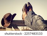Cowboy boots and hat with feet up on stables gate at ranch resting with legs crossed, country music festival live concert or line dancing concept