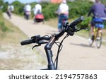 At the edge of a bike path: close-up of e-bike handlebars in the foreground, many cyclists, path and nature blurred in the background, landscape format, selective focus