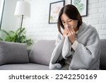 Small photo of Sick young asian woman sitting under the blanket on sofa and sneeze with tissue paper at home.