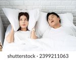 Small photo of Men snore and women cannot sleep. She uses earmuffs to muffle the snoring sound of an Asian couple in bed at home. Snoring Problems