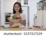 Smiling young woman holding basket with fresh vegetables standing at kitchen counter. Healthy lifestyle concept.