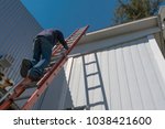 Risks of a worker climbing uninsured ladder to work on the roof of a house on a beautiful sunny day with blue sky.