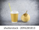 Small photo of Light brown paper disposable cup with yellow cocktail tube and white plastic lid and coconut with tubule on gray surface