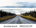 Early morning light illuminates this northern scene in the Yukon Territory of Canada. A two lane road disappears into thick fog, while above, a snowy peak sticks out above the cloud layers
