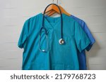 Small photo of Closeup doctors scrus with stethoscope. Green and blue surgical smock with stethoscope on wooden hanger on ceramics wall background. Closeup of doctor's scrubs with stethoscope and lab coat on hangers