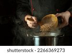 Small photo of The cook pours dry buckwheat into the pan with a spoon. Dark space for recipe or menu. The idea of cooking delicious buckwheat porridge in the kitchen.