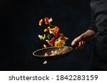 Close-up view of chef's hand throws up frying mix of colored vegetables above the pan on dark blue background. Backstage of cooking meal. Frozen motion. Food banner concept.