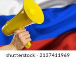 The national flag of the aggressor country Russia and a yellow megaphone in a man's hand. Russia's false propaganda, the war in Ukraine, the killing of civilians by the aggressor country.