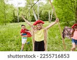 Small photo of Portrait of happy girl with tricorn hat and stick standing arms outstretched in grass by friends