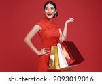 Small photo of Happy Asian shopaholic woman wearing traditional cheongsam qipao dress holding shopping bag isolated on red background. Happy Chinese new year
