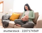 Small photo of Young asian beautiful pregnant woman hands fondle on belly sitting on sofa at home. Pregnancy, maternity, preparation and expectation concept.