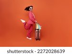 Small photo of Happy young woman traveler drag luggage and shopping bag isolated on orange copy space background, Tourist girl having cheerful holiday trip concept, Full body composition