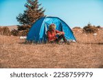Small photo of A man camping with a tent. He is surrounded by a beautiful mountain landscape, and the sun shines on his face, foreshadowing a beautiful and sunny day. The man looks satisfied and fulfilled
