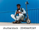 handsome African American sitting on an electric scooter, with a phone in his hands, and typing an SMS message, 5g internet, online correspondence, against background of a blue building