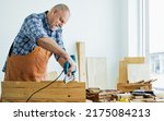 Small photo of Senior old Caucasian man wearing check shirt, apron, making DIY wooden furniture, using equipment to drill woods with happiness while standing alone at indoor home. Retirement and Hobby Concept