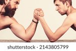 Small photo of Heavily muscled bearded man arm wrestling a puny weak man. Arms wrestling thin hand, big strong arm in studio.