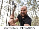 Small photo of Brutal bearded guy in a black T-shirt in a summer forest with an ax in his hands. Manifestation of negative emotions. Threateningly brandishes an ax and shouts. Natural landscape