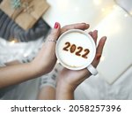 Small photo of Number 2022 on frothy surface of cappuccino served in white cup holding by female hands on blurred white bed with string lights. New year new you, Holidays food art theme Happy New Year. (top view)