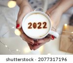 Number 2022 on frothy surface of cappuccino served in white cup holding by female hands on blurred white bed with string lights. New year new you, Holidays food art theme Happy New Year. (top view)