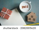 Small photo of White coffee cup with number 2022 on frothy surface flat lay over green background with house model, gift box, yellow pen and blank notebook paper. Happy new year 2022 theme. (close up, top view)