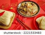 Small photo of Chinese Lunar New Year lucky food longevity noodle or stir fried noodles served on red background with red envelopes, wooden chopsticks, ancient gold bullion nugget and uncooked dried noodles.