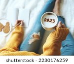 New Year's goal setting, number 2023 on frothy surface of cappuccino in white coffee cup holding by woman in yellow knitted sweater with jeans sitting on bed while writing down her resolutions.