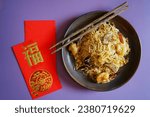 Small photo of Longevity noodles or stir fried noodle in dark plate with chopsticks on purple background with blurred red envelope with blessing word means ‘good fortune’ Chinese Lunar New Year lucky food (top view)