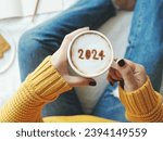 2024 new year's goal setting, number 2024 on frothy surface of cappuccino in white coffee cup holding by woman in yellow knitted sweater with jeans sitting on bed with white blanket background.