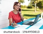 Woman working on modern laptop outdoor against a white wooden background - 40 years old woman calling while taking notes - Freelancer or female entrepreneur working with her devices from remote