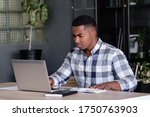 Small photo of Serious young african american freelancer works remotely at home using a laptop high speed internet sitting at the table. Young mixed race businessman working after hours