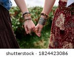 Close-up picture of hands with colorful bracelets and various rings, holding each other. Hippie women, wearing boho style clothes, standing on green field. Female friendship concept.
