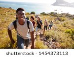 Small photo of Millennial African American man leading friends hiking single file uphill on a path by the coast